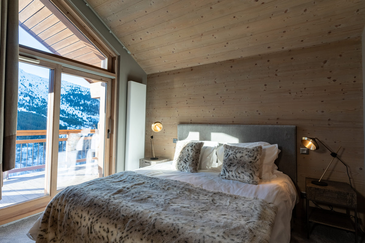 A cozy bedroom with a neatly made bed featuring patterned pillows and a textured throw blanket, set against a wood-paneled wall. Large windows on the left offer a view of snowy mountains outside.