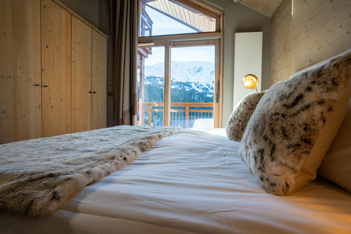 A cozy bedroom scene with a neatly made bed in the foreground, white bedding, textured beige and brown pillows, and a large window looking out onto snowy mountains. The room has light wood walls, a wooden wardrobe, and a small wall lamp.