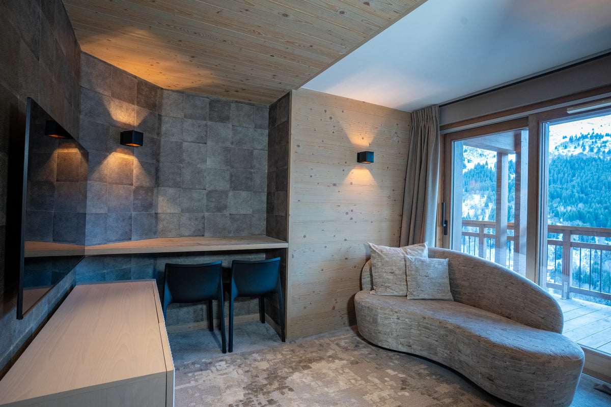 A luxury chalet-style living room featuring a curved upholstered sofa, a built-in desk with two dark chairs, a stone-tile accent wall, and large glass doors looking out to a forested mountain view.