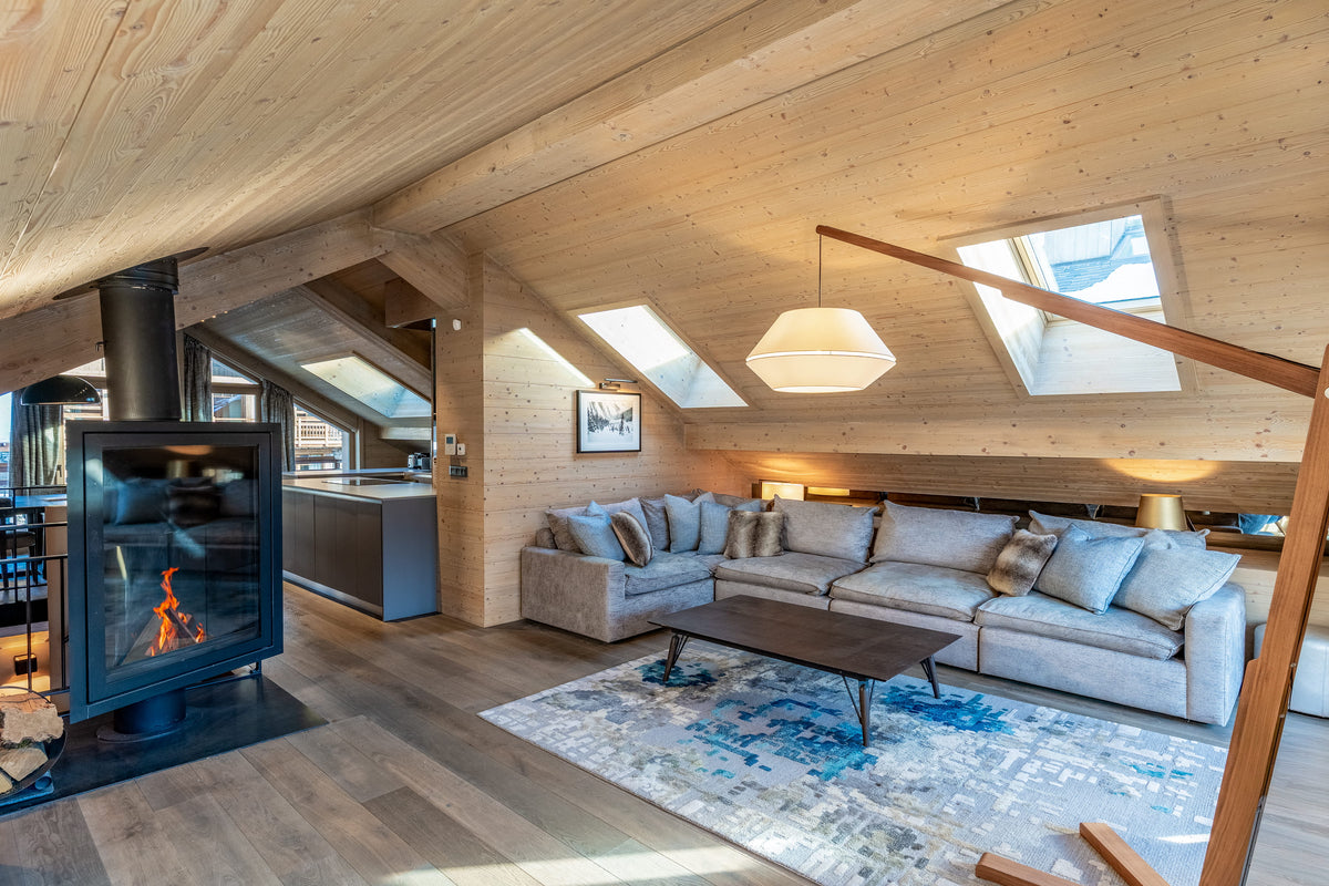 A modern ski chalet living room featuring a light wood paneled ceiling and walls, a gray sectional sofa, a dark wood coffee table, a blue patterned area rug, and a black wood-burning stove.