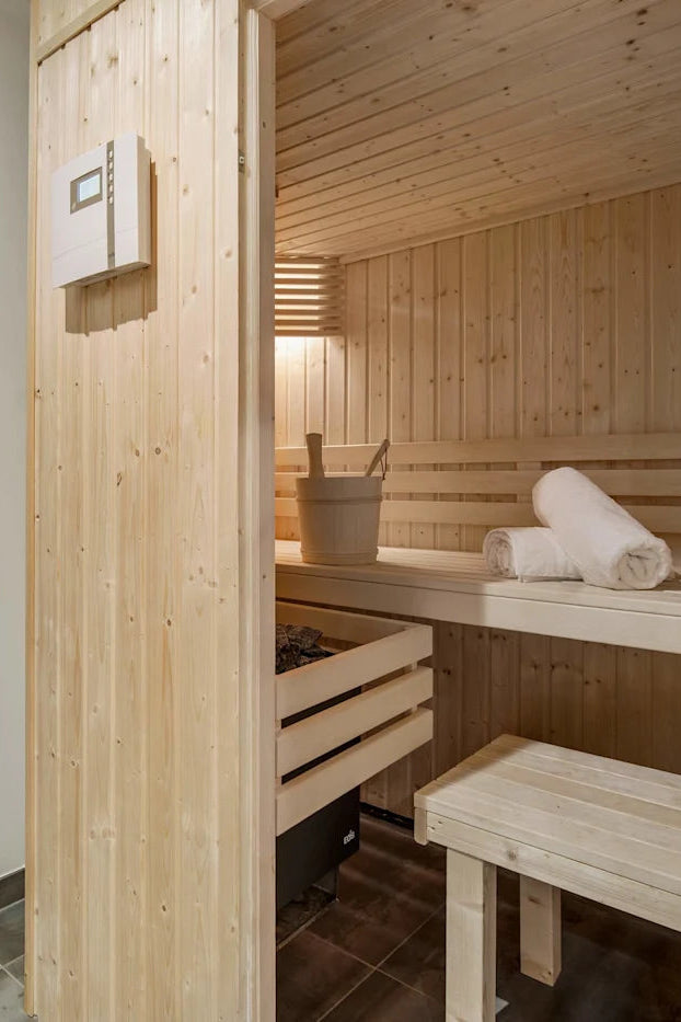 A modern sauna and shower cabin interior with light wood walls and benches, a tiled shower area, and rolled towels on the bench.