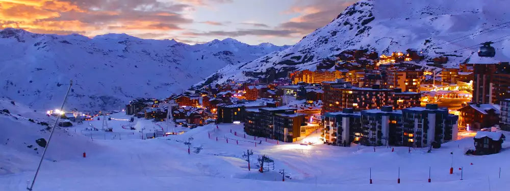 Snow-covered mountain village at dusk with illuminated buildings, ski slopes in the foreground, and a dramatic orange and blue sunset sky.