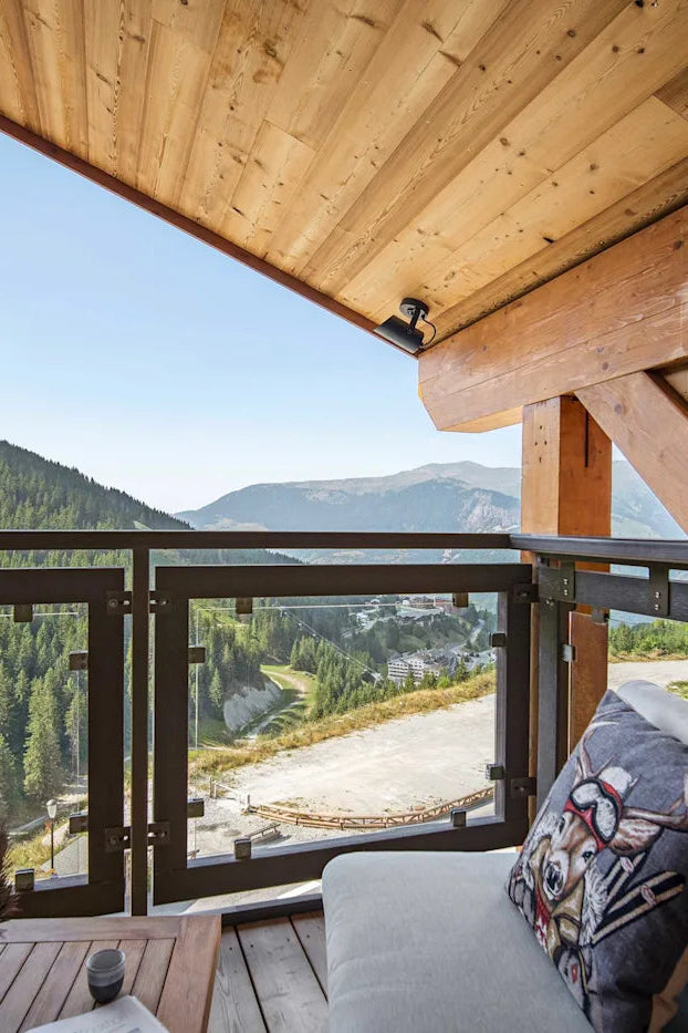 A modern wooden chalet balcony with a gray outdoor sofa and decorative pillow, overlooking a lush green mountain valley under a clear blue sky.