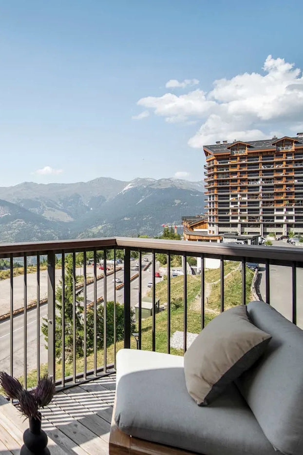 A modern outdoor balcony with a wooden railing and light gray lounge chair, overlooking a scenic mountain road and forested slopes under a blue sky. A tall resort-style building is visible on the right.