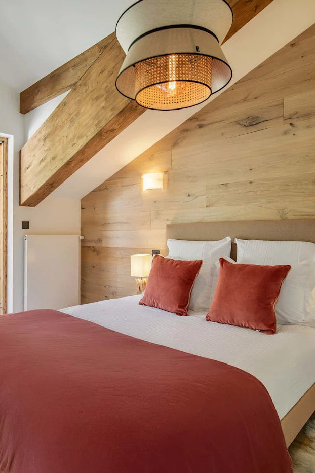 A cozy bedroom interior with a neatly made bed featuring white linens and rust-red pillows and blanket, set against a light wood-paneled wall. Large glass doors open onto a balcony with a grassy outdoor view, and warm wall lights and a hanging pendant lamp illuminate the space.