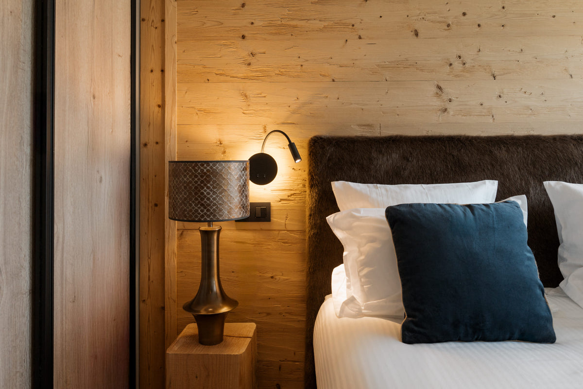 A cozy bedroom corner with a bed dressed in white linens and a dark blue accent pillow, set against a light wood-paneled wall with a lit wall sconce and a patterned table lamp on a wooden bedside table.
