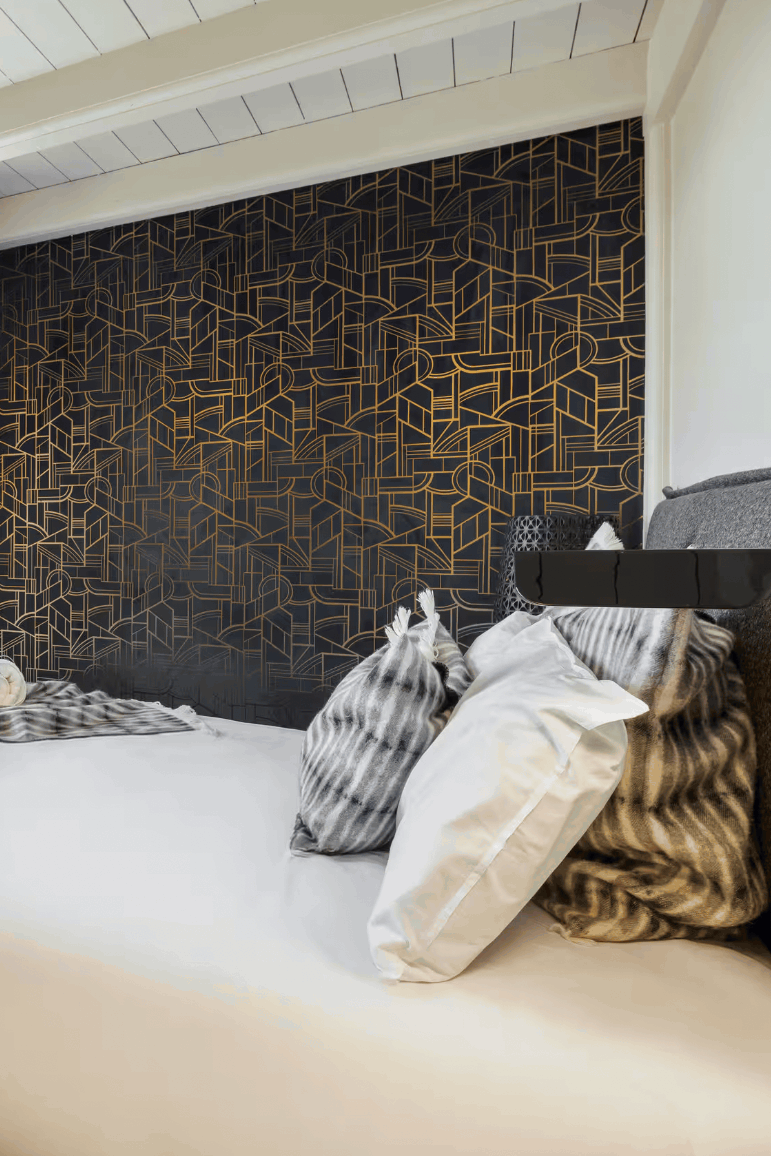 A modern bedroom interior featuring a neatly made bed with white linens, striped pillows, and a striped throw blanket, set against a dark patterned accent wall. Large windows with dark curtains offer a view of snowy mountains outside.