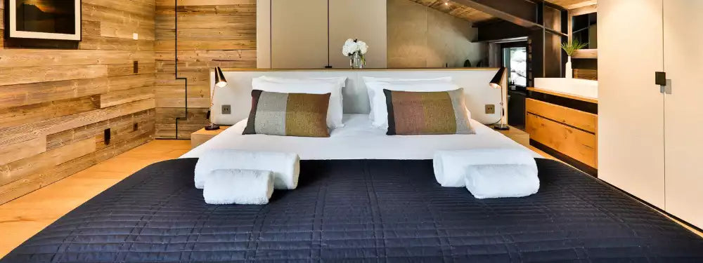 A modern bedroom interior featuring a large bed dressed in white linens and folded white towels, set on a dark quilted bedspread. The room has wood-paneled walls, a wood vanity, and a built-in kitchen area in the background.