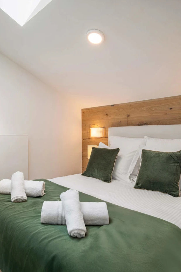 A neatly made bed in a modern bedroom with white linens, dark green pillows and throw, a wooden headboard, bedside lamps, and rolled towels on the bedding.