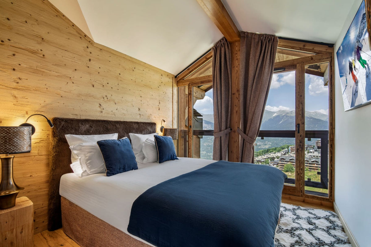 A modern mountain-view bedroom with a large bed dressed in white linens and a navy blue throw, set against a light wood-paneled wall with dark brown curtains and floor-to-ceiling windows overlooking a scenic alpine landscape.