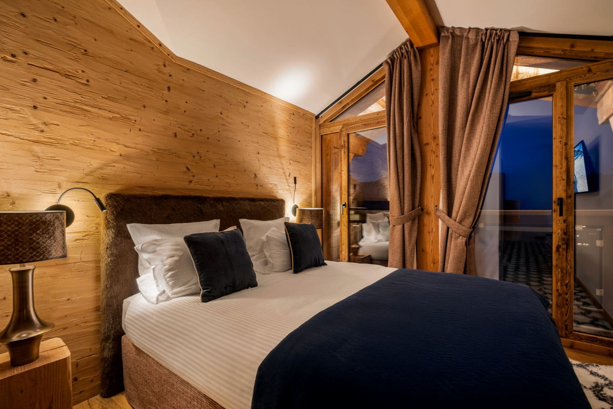 A cozy mountain-style bedroom with a neatly made bed, white bedding, dark navy throw blanket, and dark accent pillows. The room features warm wood-paneled walls, a wooden headboard, bedside lamps, and large curtains framing a glass door that looks out onto a balcony at night.