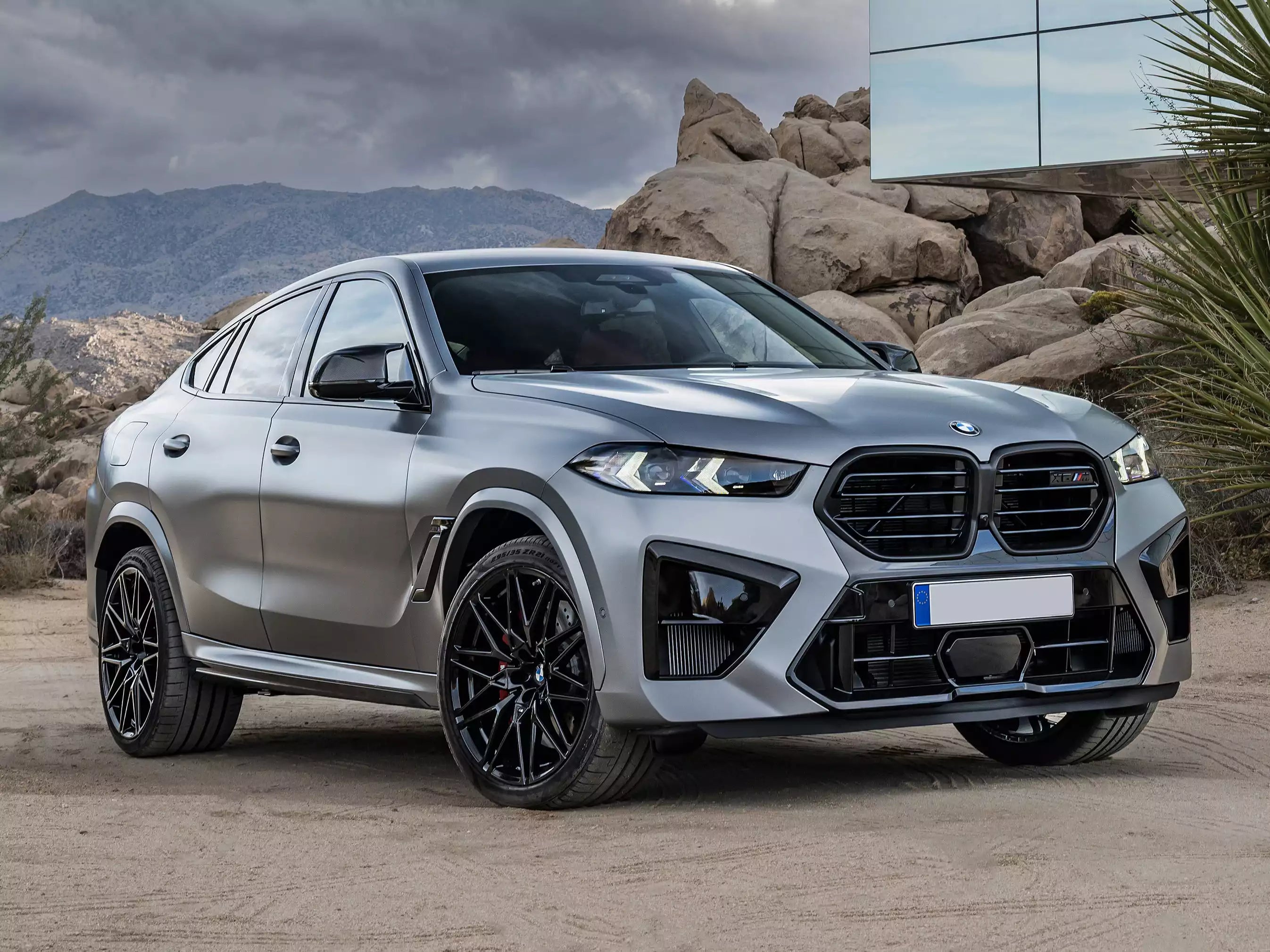 A silver BMW X6 M is parked outdoors on a dirt surface with rocks, mountains, and a modern glass building in the background. The car is shown at a three-quarter front angle, featuring its distinctive M grille, black wheels, and sleek body lines.