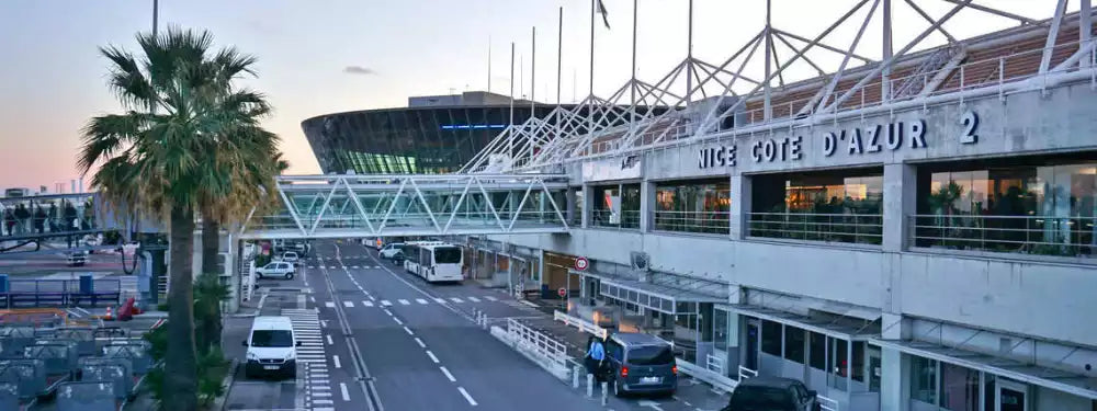 A wide outdoor shot of the Nice Côte d'Azur 2 airport terminal building at dusk, with a road running through the foreground, palm trees on the left, and vehicles in front of the terminal.