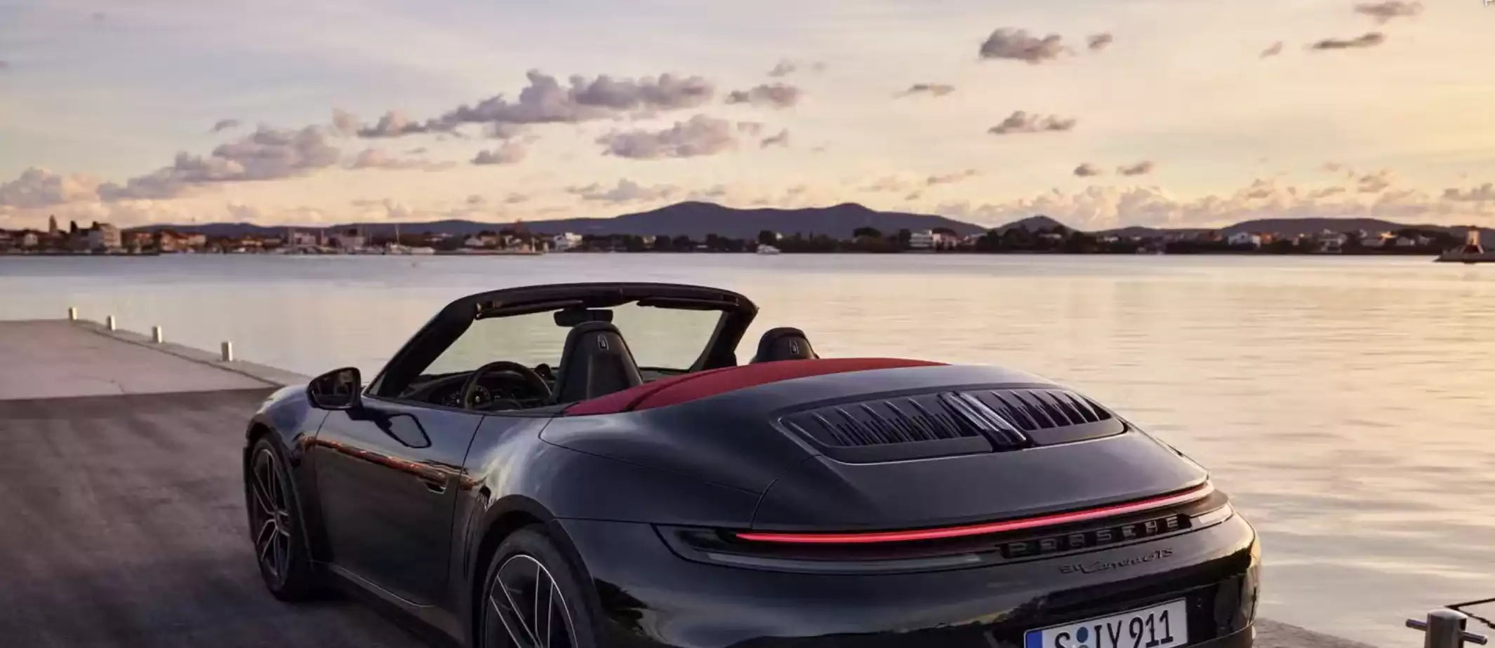 A dark gray Porsche 911 Turbo S convertible with a red interior roof lining is parked on a waterfront promenade at sunset.