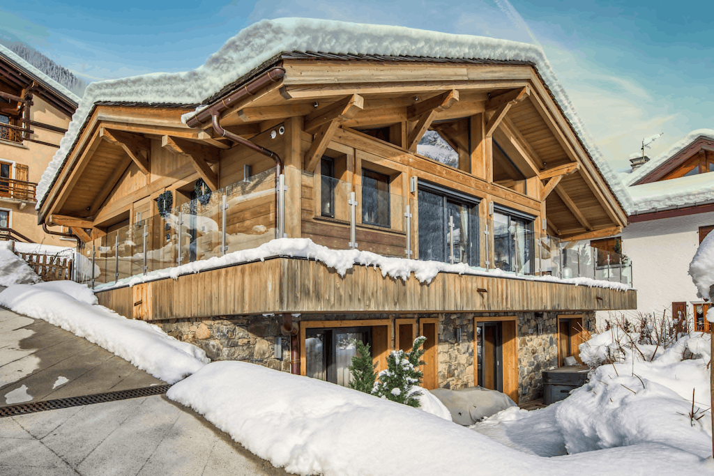 A large alpine chalet-style wooden house with snow-covered roofs and balconies, surrounded by deep snowbanks and outdoor seating areas in a winter mountain setting.
