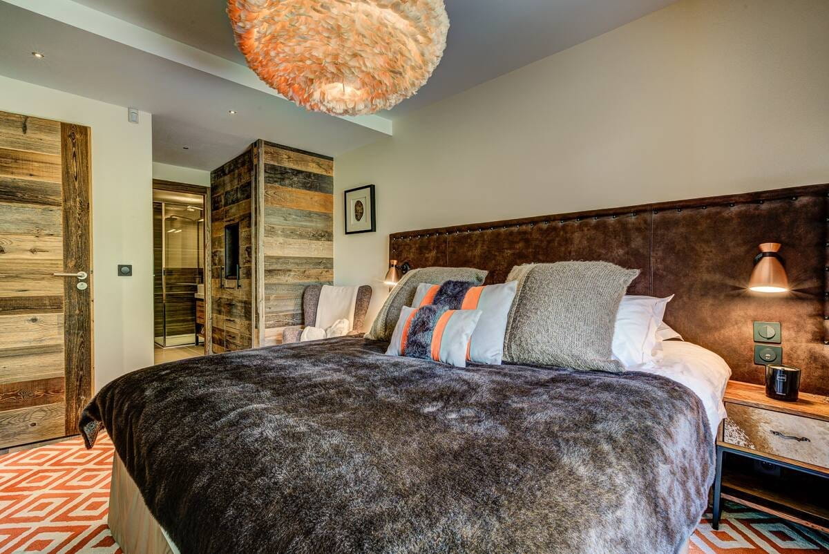 A rustic-style bedroom featuring a large bed with a textured brown faux-fur duvet, a dark brown leather headboard, and a mix of gray, blue, and orange accent pillows. The room has warm beige walls, a large woven pendant light, and a geometric orange and white rug.