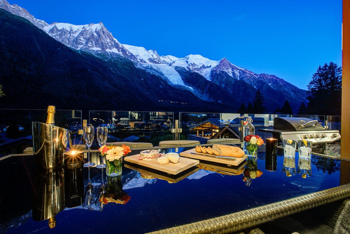 A luxury outdoor dining setup on a glass-enclosed balcony overlooking a mountain village at dusk, with snow-capped peaks under a deep blue sky. The table is styled with an open bottle, two champagne flutes, candles, small flower arrangements, cheese on wooden boards, and a stainless steel grill.