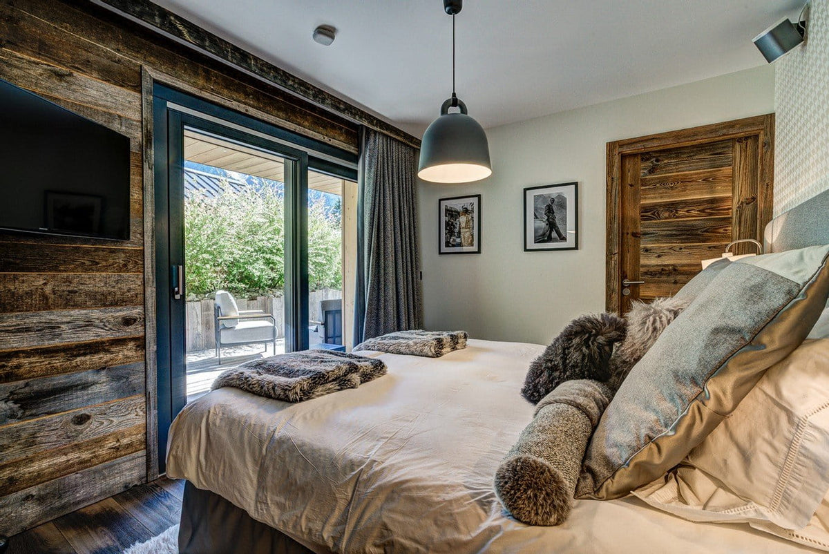 A cozy bedroom interior featuring a large bed with beige bedding, textured gray and brown pillows, rustic wood-paneled walls, a hanging pendant light, and sliding glass doors leading to a patio.