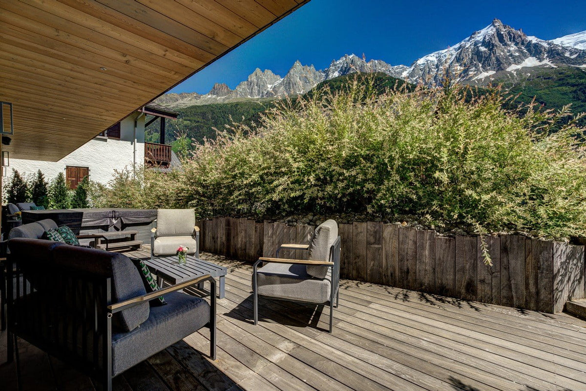 Outdoor patio furniture set on a wooden deck with a mountain view. The furniture includes a dark gray sectional sofa, a matching armchair, and a light gray lounge chair with a coffee table.