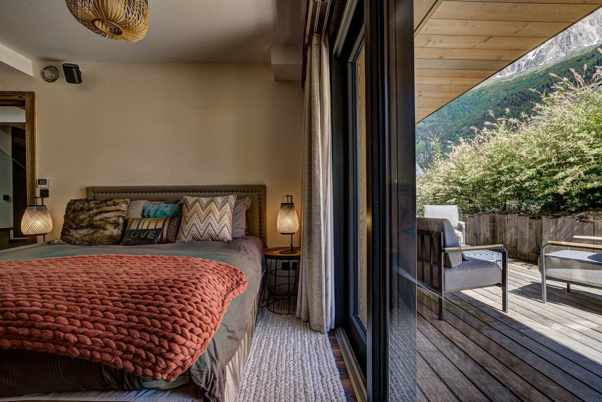 A cozy bedroom interior with a bed dressed in gray bedding and a chunky rust-red knit throw, set beside large sliding glass doors that open onto a wooden deck with outdoor seating and a mountain landscape in the background.