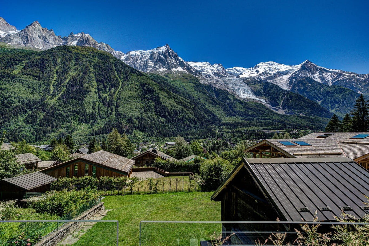 A scenic mountain landscape with snow-capped peaks under a deep blue sky, overlooking green valley homes and grassy lawns in the foreground.