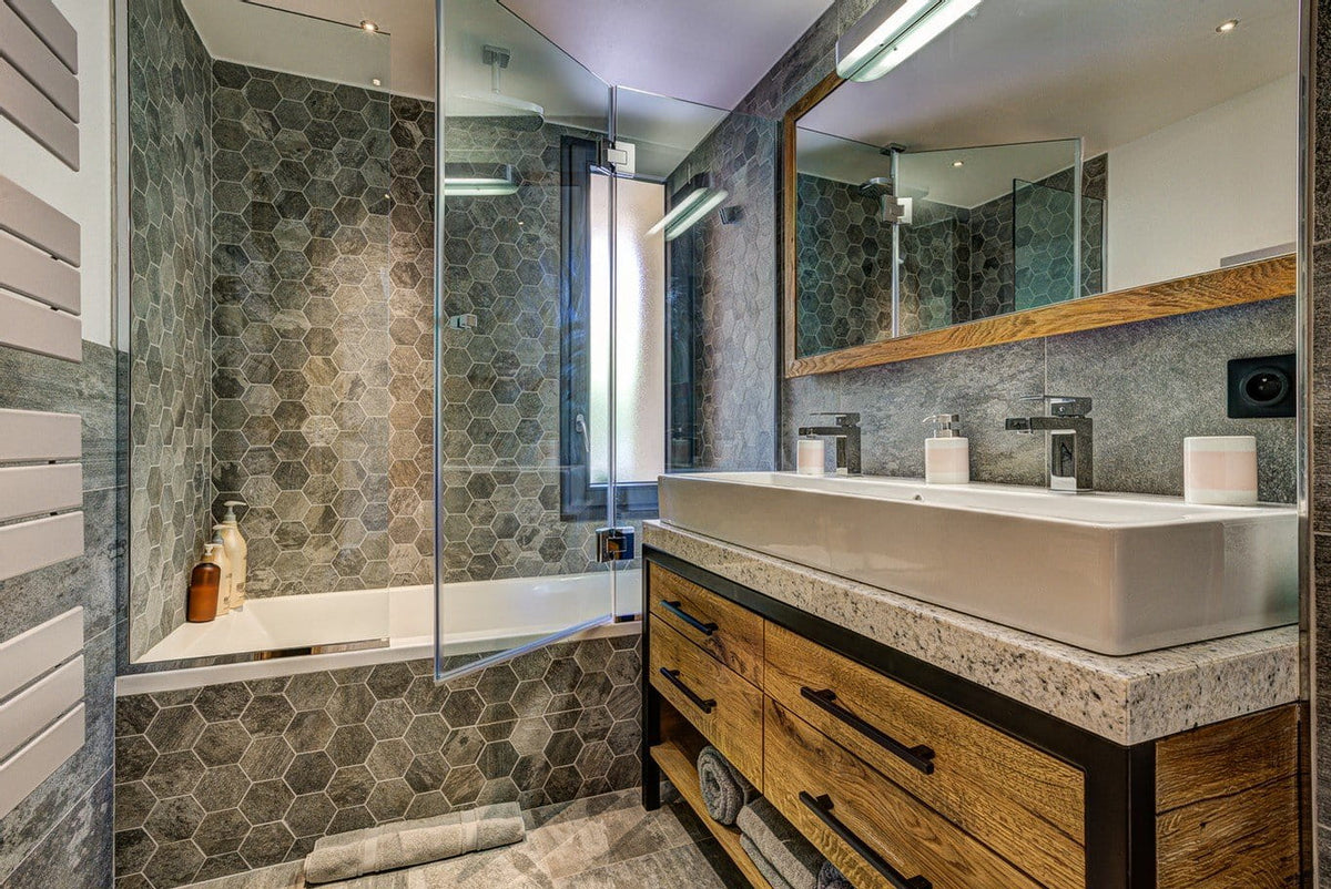Modern bathroom vanity with a white rectangular vessel sink, chrome faucets, a large mirror, and a wood-and-metal cabinet. A glass shower enclosure and hexagonal gray tile wall are visible, along with a bathtub and stone countertop.