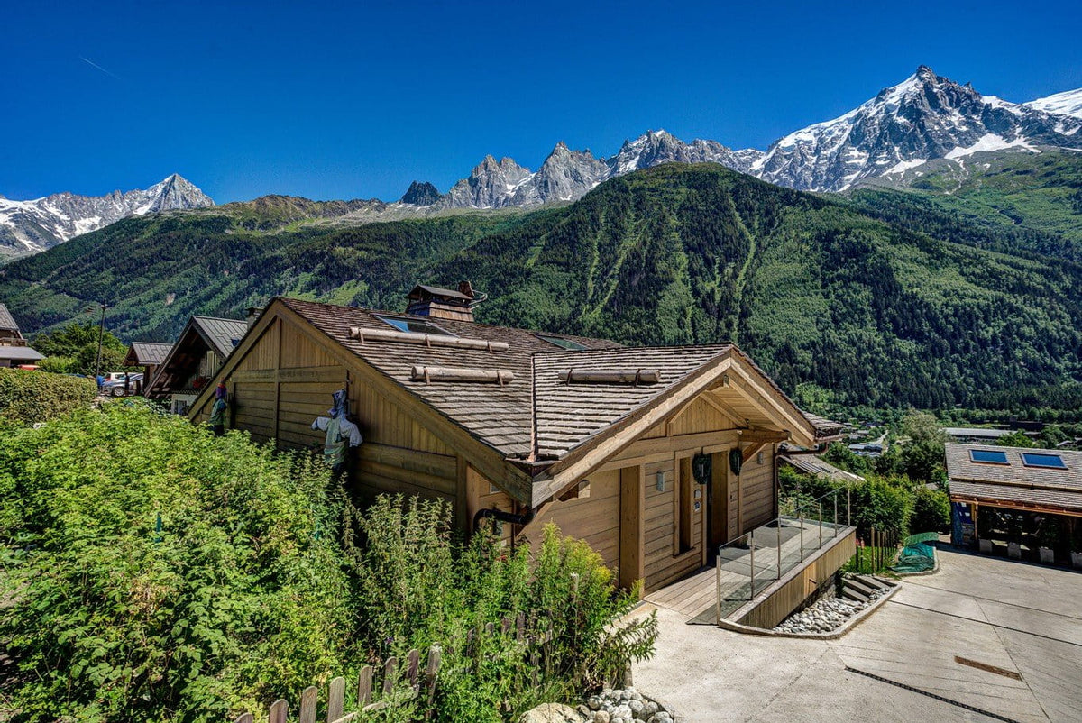 A wooden chalet-style mountain house with a tiled roof, situated in a lush green alpine setting with snow-capped peaks under a clear blue sky.
