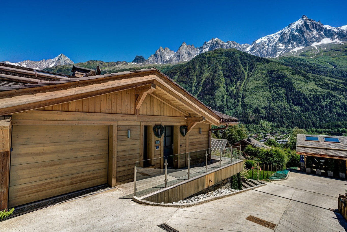 A modern wooden chalet-style building with a large garage door and small porch, set against a dramatic mountain landscape with snow-capped peaks and green slopes under a clear blue sky.