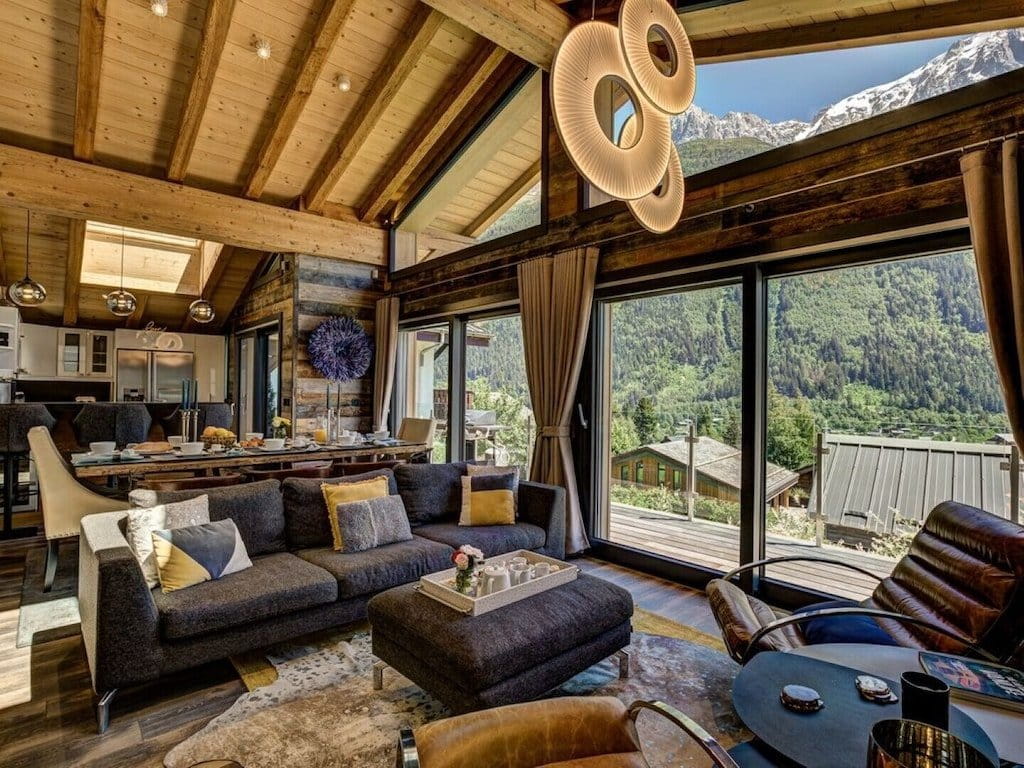 A luxury modern chalet living room with a dark gray sectional sofa, wooden ceiling beams, and large windows overlooking a lush green mountain valley.