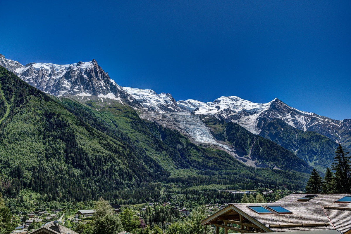 A scenic mountain landscape with snow-capped peaks, deep green forested slopes, a valley below, and rooftops in the foreground under a clear blue sky.