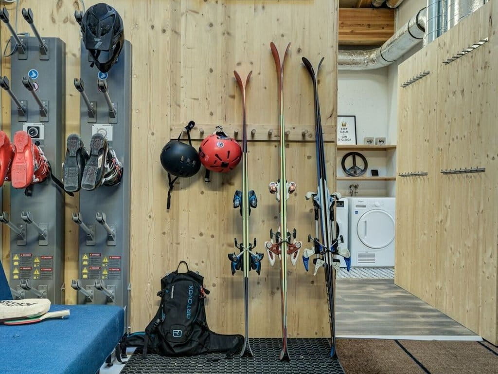 A ski boot storage rack in a rustic wooden lodge setting, holding various skis, helmets, and boots, with a blue bench in the foreground and a laundry room visible in the background.