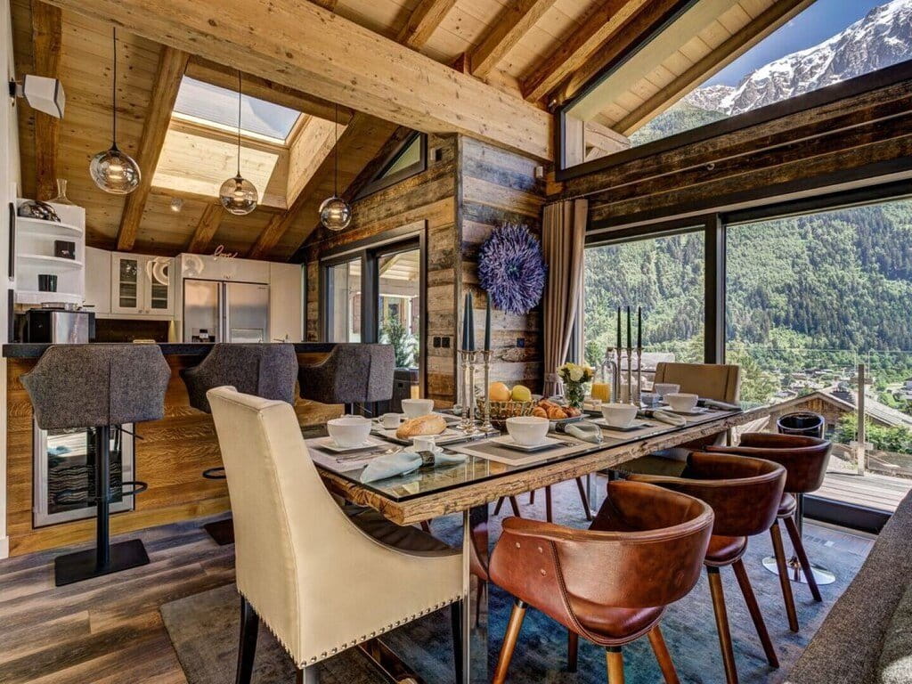 A luxury alpine chalet dining room featuring a long wooden dining table set with white dishes, surrounded by brown leather and cream upholstered chairs, with a kitchen and stunning mountain views through large windows.