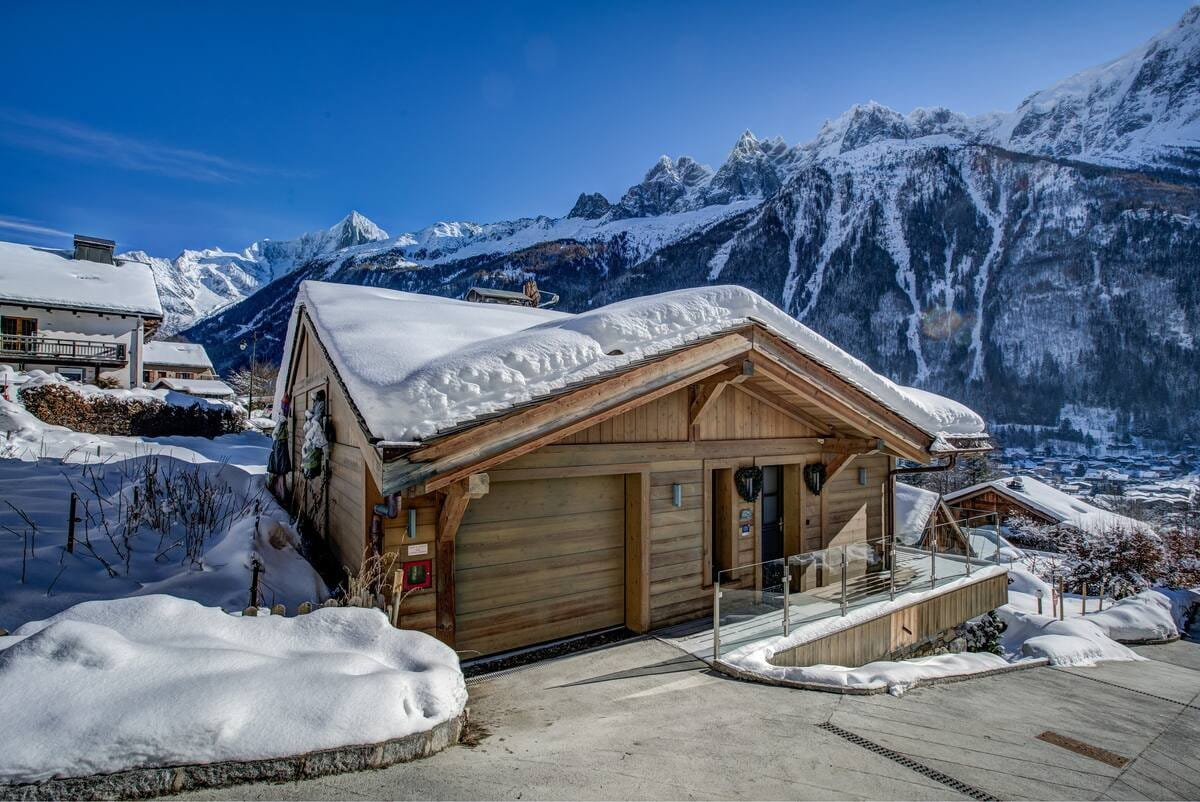 A snow-covered wooden chalet with a large garage door, set against a backdrop of snow-dusted mountains and a clear blue sky.