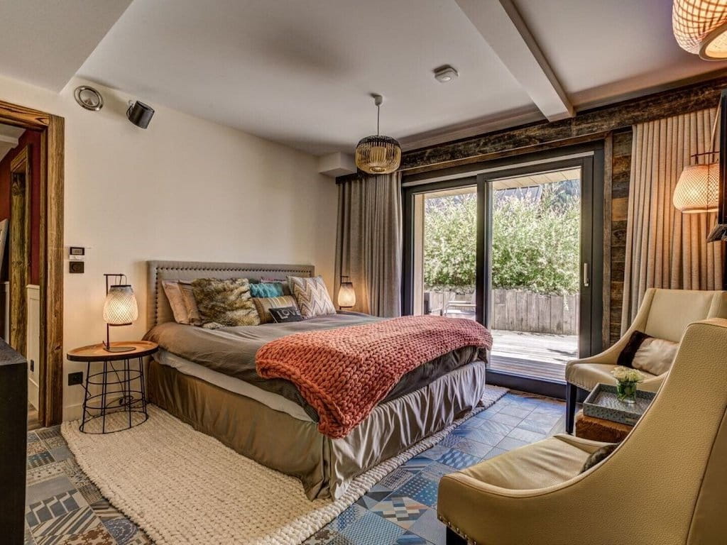 A cozy bedroom interior featuring a bed with a textured rust-red throw blanket, neutral bedding, a patterned rug, a beige armchair, and large windows looking out to a garden patio.