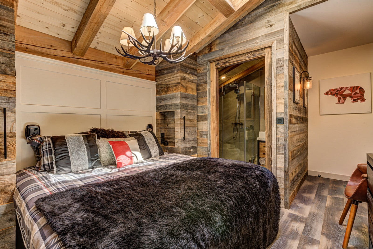 A rustic bedroom interior with a large bed dressed in plaid bedding and a dark faux-fur throw blanket, set in a cabin-style room with wood-paneled walls, a chandelier, and a glass shower enclosure in the background.