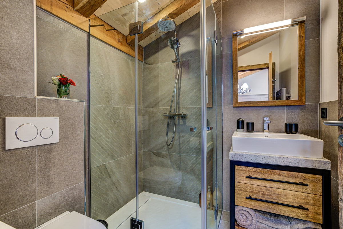 Modern bathroom interior with a glass shower enclosure, gray stone-look tiles, a white sink basin, wood-look vanity cabinet, and a wall-mounted mirror.
