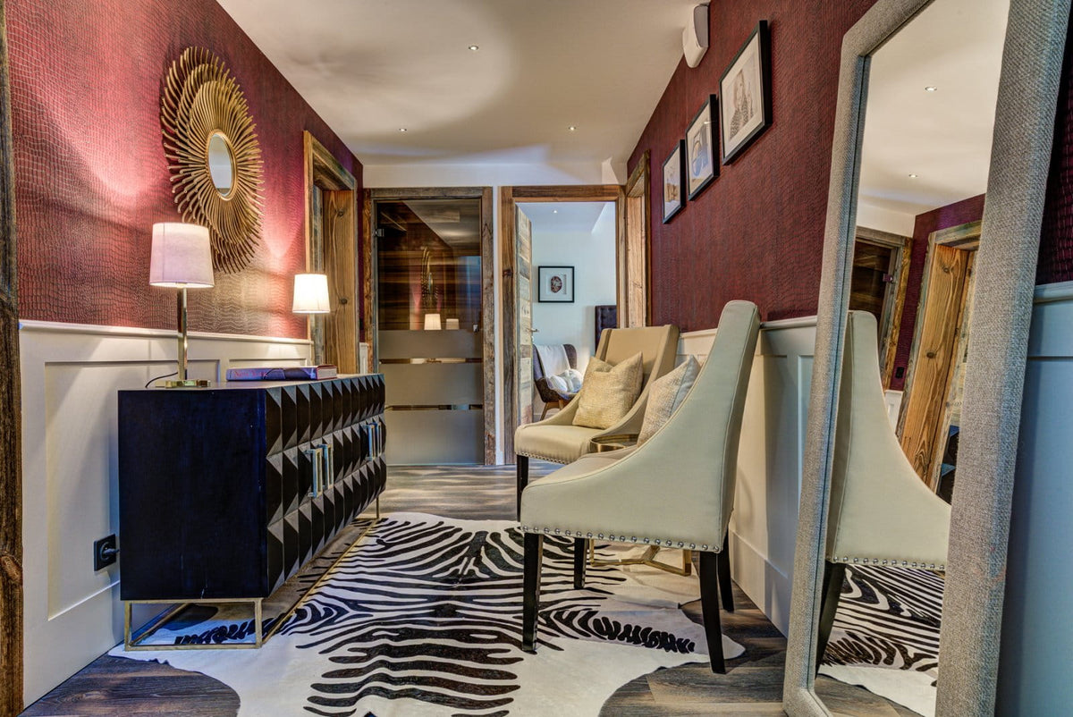 A styled interior hallway featuring a black tufted sideboard with lit table lamps, a cream upholstered chair, a zebra-print rug, and deep burgundy textured walls with framed art.