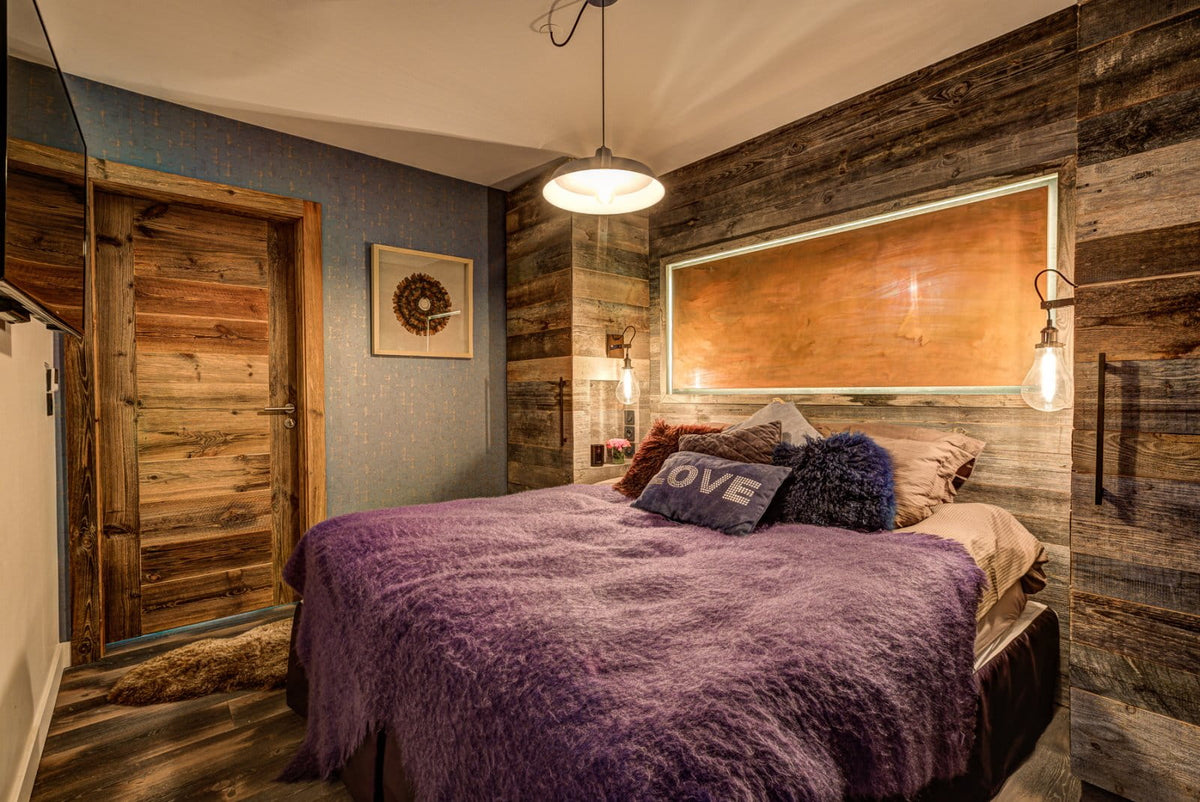 A rustic bedroom featuring a bed with a textured purple shag blanket and pillows, set against a wall made of dark wood planks with an integrated light. A blue accent wall and a wooden door are visible on the left.