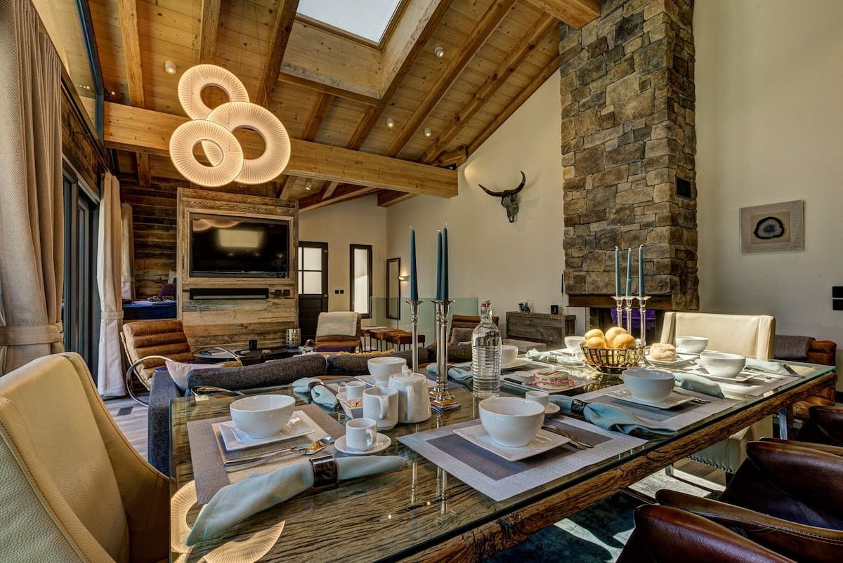 A luxury dining room with a rustic wooden table set for a meal, surrounded by cream-colored chairs. The room features a high wood-beamed ceiling, a stone fireplace, and a modern living area in the background.