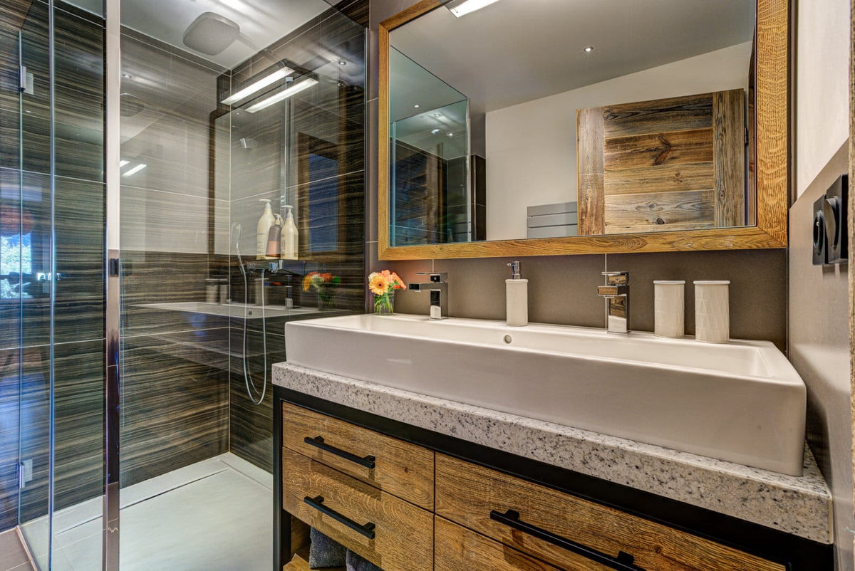 Modern bathroom vanity with a white rectangular vessel sink, wood-look cabinets, a speckled stone countertop, and a large wood-framed mirror above. A glass shower enclosure is visible to the left.