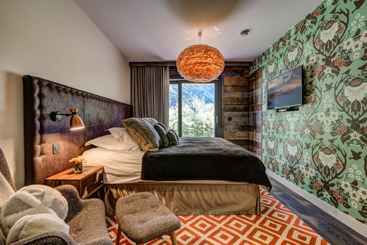 A styled mountain-view bedroom featuring a bed with white linens and dark bedding, a large tufted headboard, patterned wallpaper, a hanging woven pendant light, and an orange geometric rug.