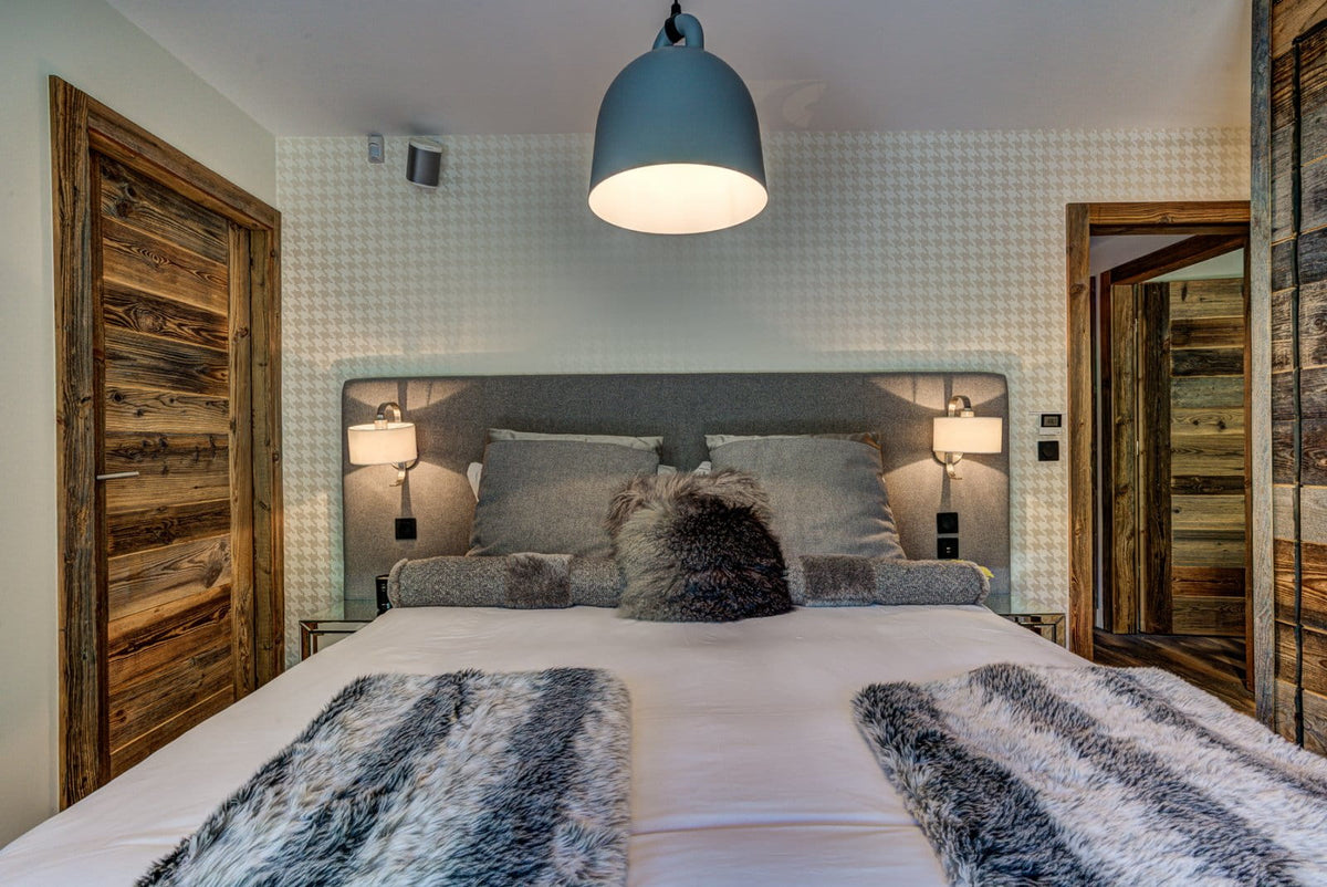A modern bedroom featuring a large bed with white linens, gray pillows, and textured gray and black faux fur throws. The bed is set against a white patterned wall with a gray upholstered headboard, flanked by bedside tables with lamps.