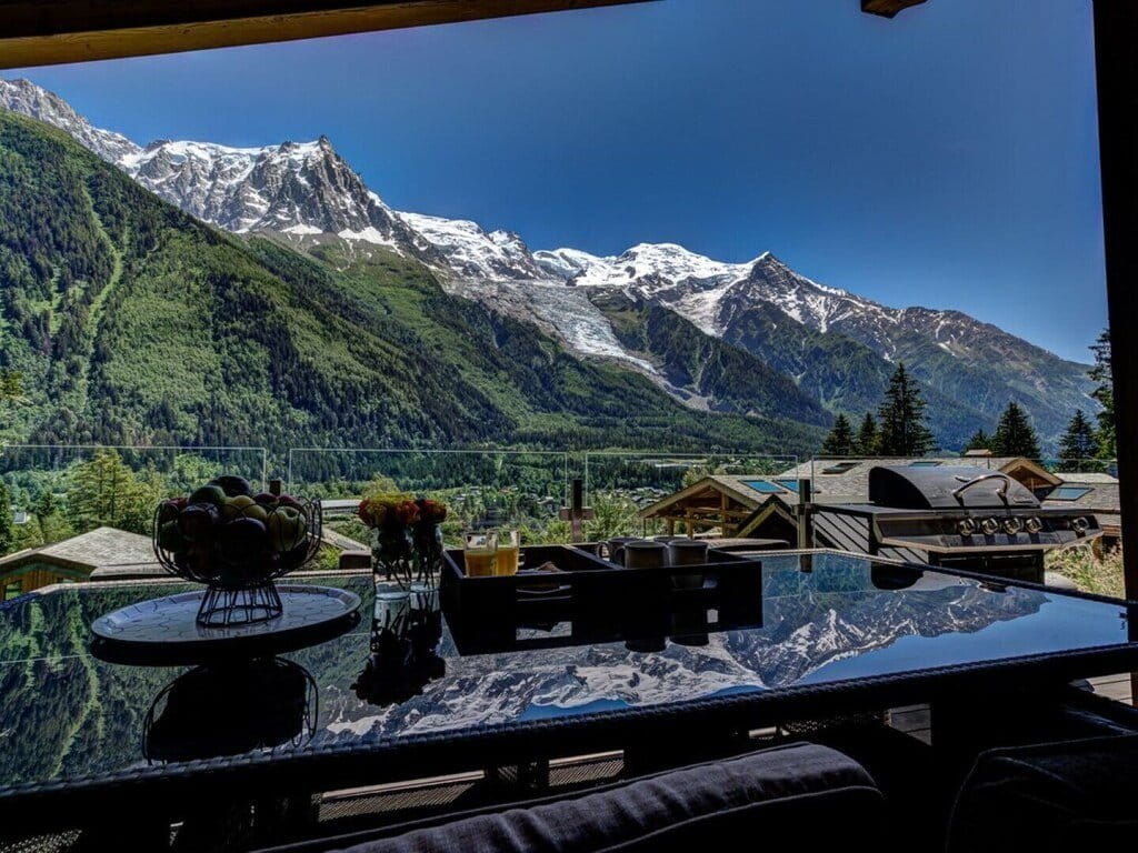 A scenic view of snow-capped mountains and green valleys from a high-rise balcony or terrace, with a glass table in the foreground reflecting the landscape and a few dining items.