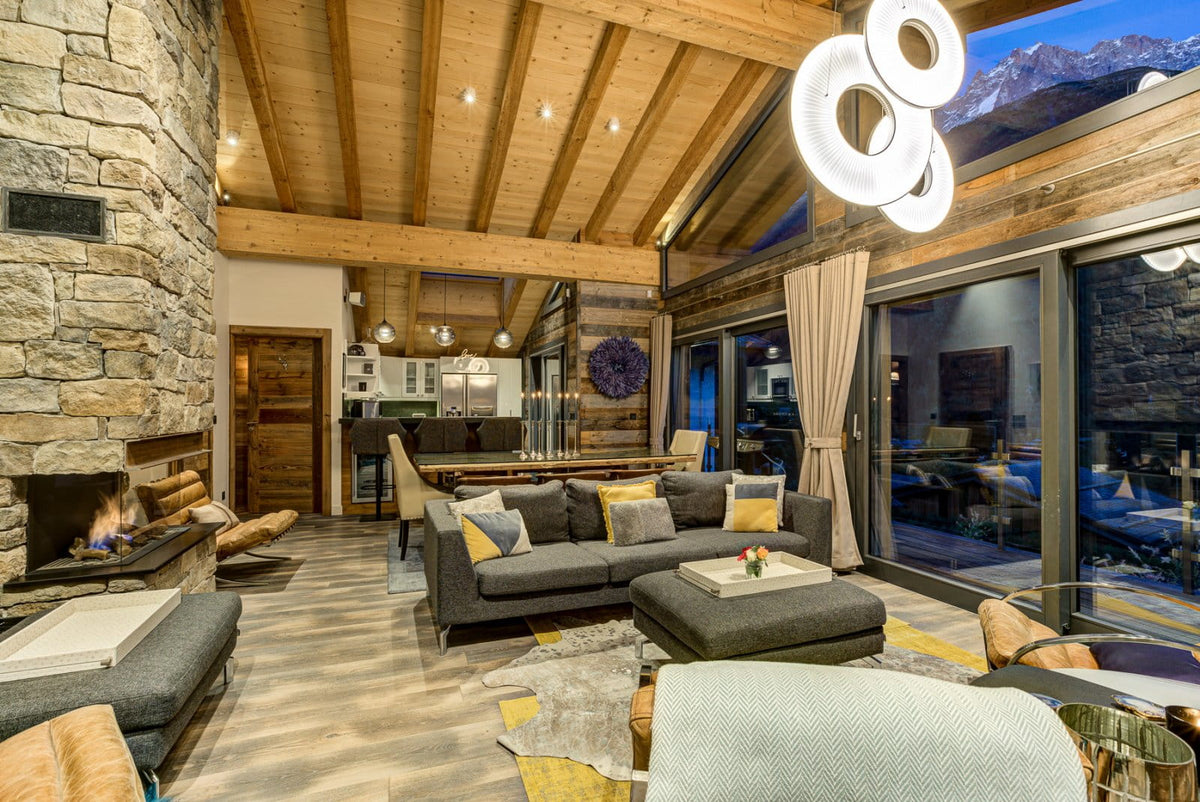 A luxury modern chalet living room featuring a dark gray sectional sofa with yellow accent pillows, a stone fireplace, a high wood-beamed ceiling, and large windows looking out onto a mountain view.
