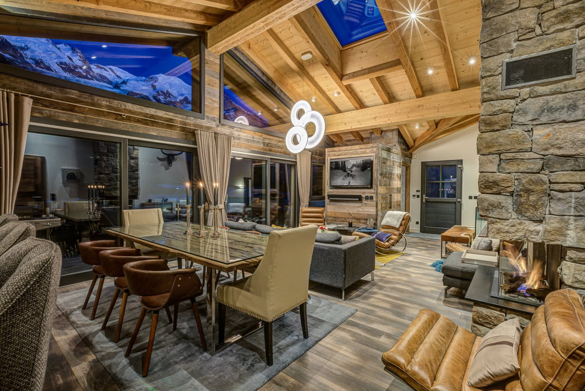 A luxury chalet-style mountain lodge interior with a dining table, beige chairs, stone walls, a fireplace, wood ceiling, and large windows showing a snowy mountain view.