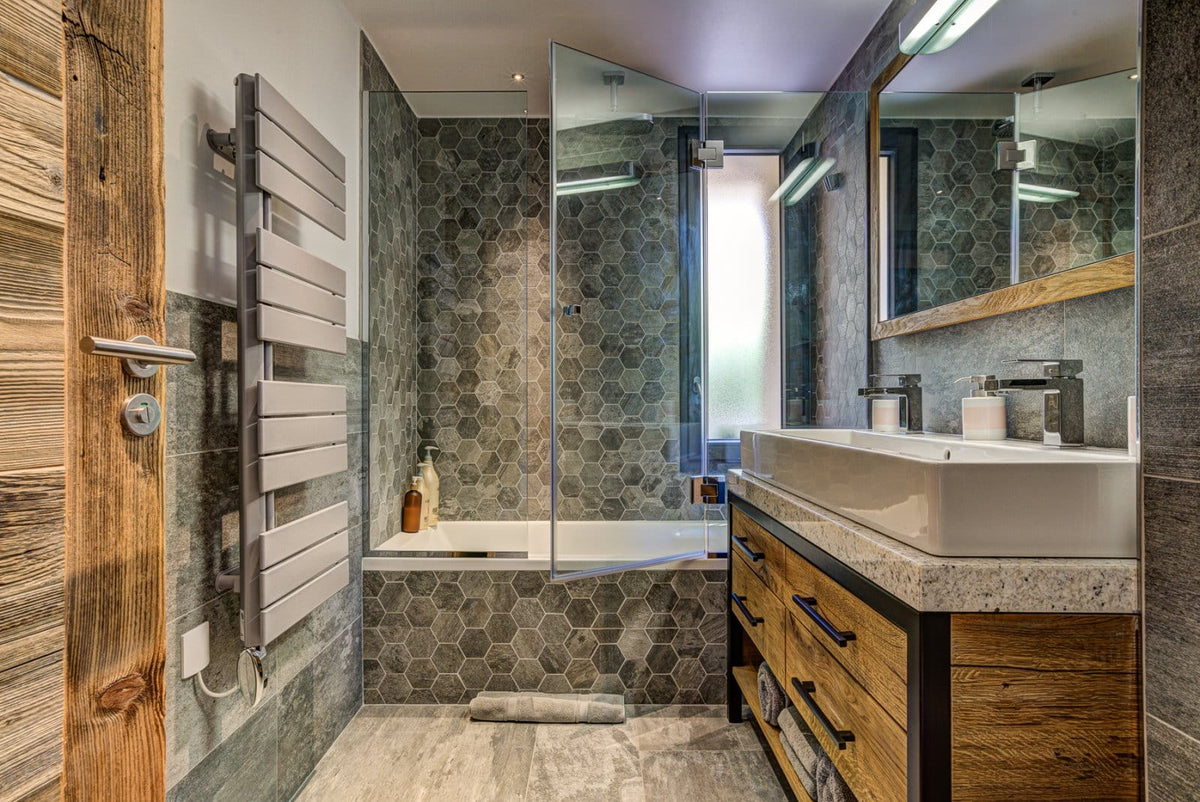 A modern bathroom interior with a white vessel sink, wood vanity, gray hexagonal wall tiles, a glass shower enclosure, and a tall wood radiator heater.