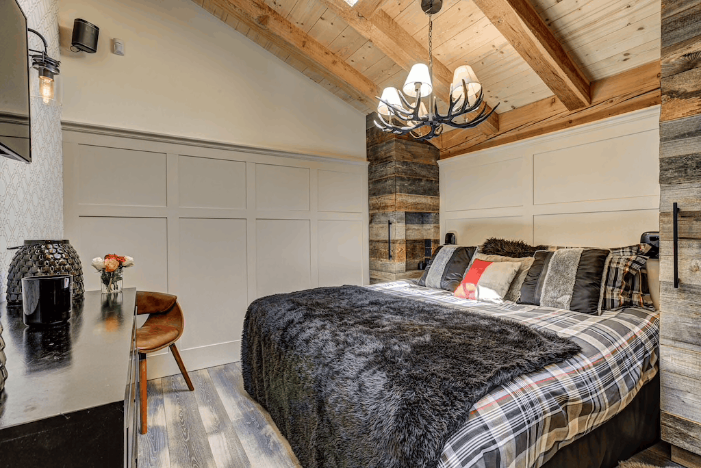 A rustic bedroom interior with a large bed dressed in gray and plaid bedding, set under a wood-beamed ceiling with a hanging chandelier. White paneled walls, a stone accent column, and a dark vanity table with a chair are visible.