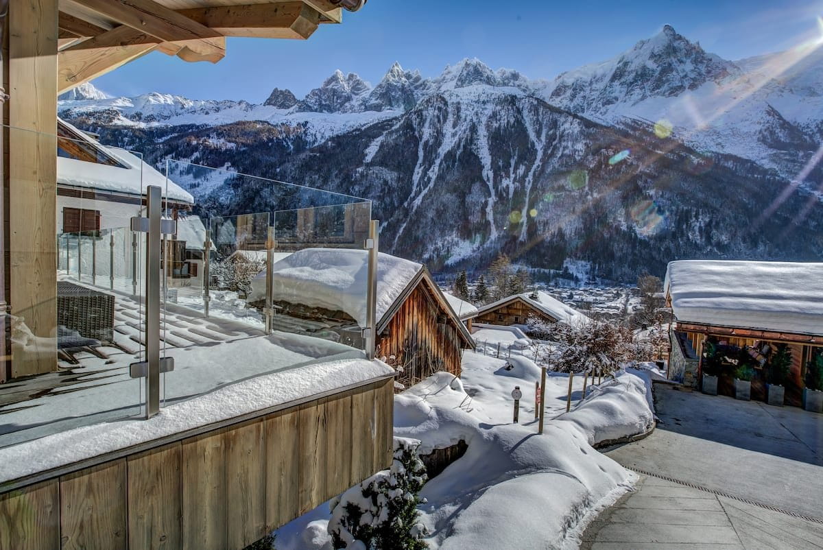 A sunny winter mountain village scene with snow-covered chalets, wooden balconies, and dramatic snow-dusted peaks under a clear blue sky. No people or animals are visible.