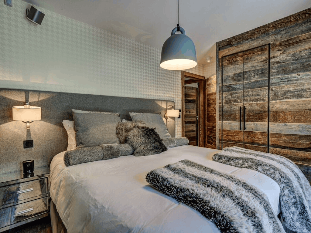 A modern bedroom featuring a bed dressed in white bedding with gray and black textured pillows and throws, set against a gray upholstered headboard. The room includes a rustic wood-paneled wardrobe, a textured light gray wall, bedside tables with lamps, and a hanging pendant light.