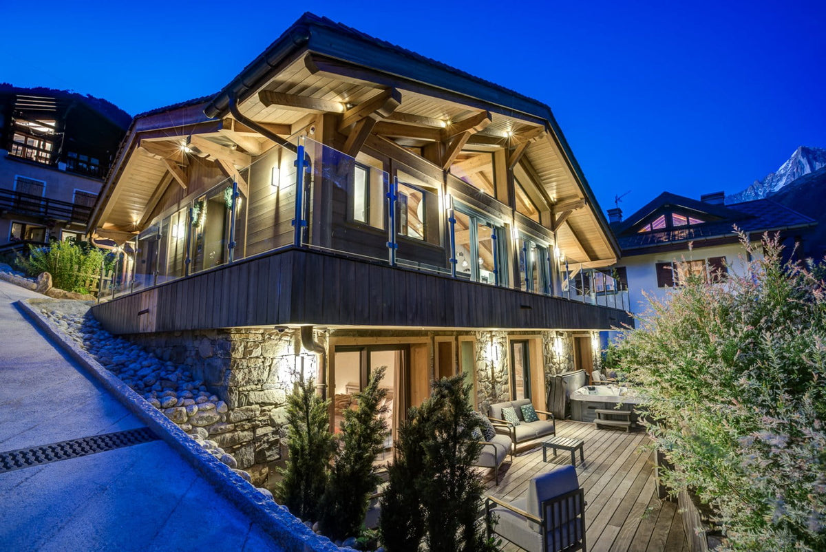 A luxury chalet-style mountain lodge at dusk, featuring warm wood and stone exterior walls, illuminated windows, a large wooden deck with outdoor seating, and snow-covered slopes in the background under a deep blue twilight sky.